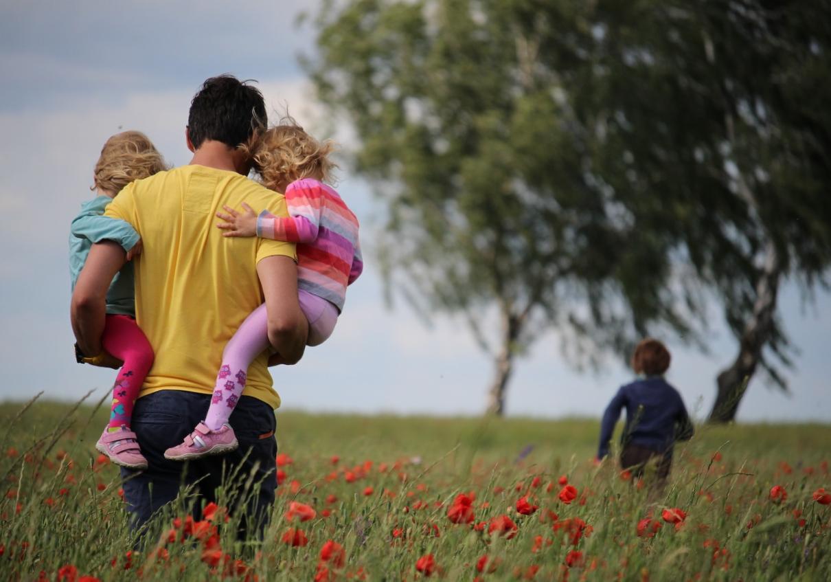 Man holding twin daughters and watching his son run through a field